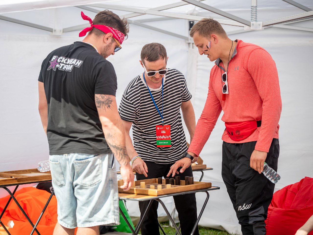 Три участника играют в деревянную настольную игру под шатром на мероприятии: игроки сосредоточены, у одного бейдж, летняя фестивальная атмосфера вокруг.