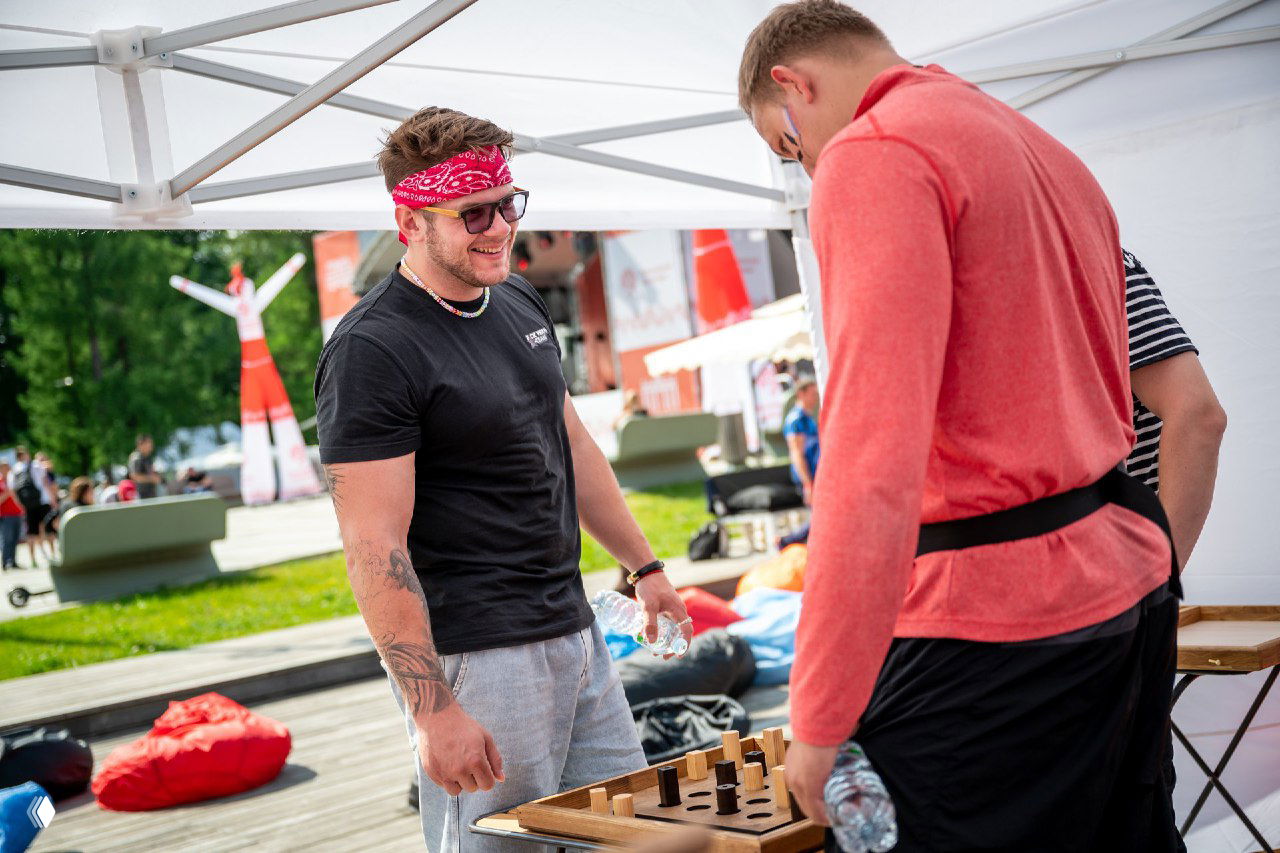 Группа людей у столика с крафтовыми деревянными играми на уличном событии: улыбки, неформальная одежда и фон с зеленью и пуфами создают дружелюбную обстановку.