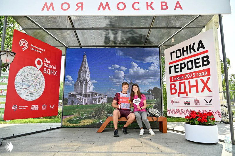 Двое людей сидят на скамейке перед крупным светодиодным экраном и рекламными баннерами «Гонка Героев» на ВДНХ — пример арендуемого LED-решения.