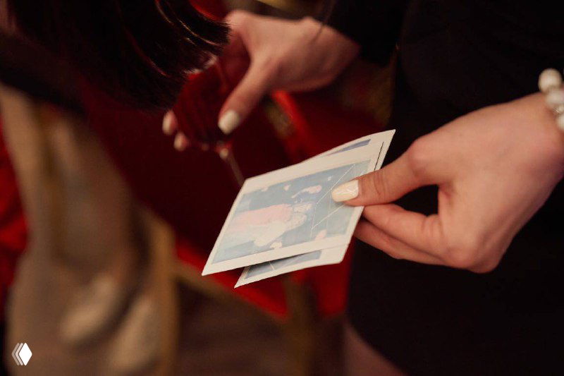 Руки держат только что распечатанную моментальную фотографию формата Instax/Polaroid; видно бумажную фотографию и детали рук гостей на мероприятии.