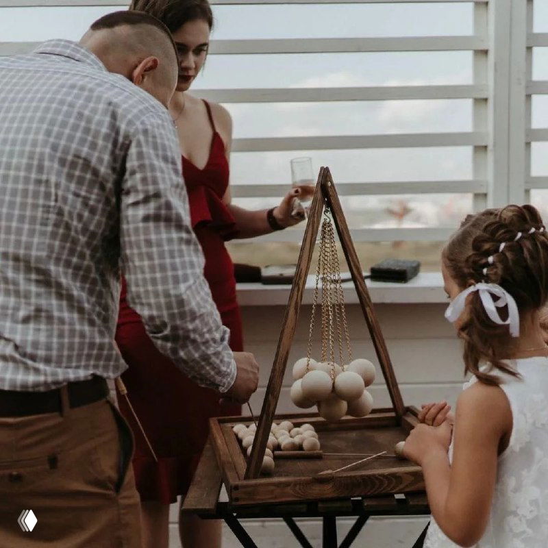 Гости мероприятия — взрослые и ребёнок — играют в крафтовую деревянную настольную игру с треугольной рамой и шариками на террасе события.