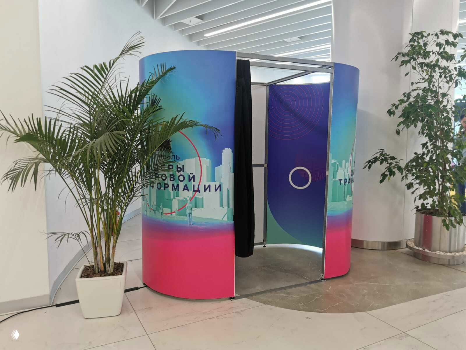 Классическая круглая фотобудка с чёрной шторкой и ярким градиентным брендированием, установлена в светлом холле рядом с горшечными растениями.