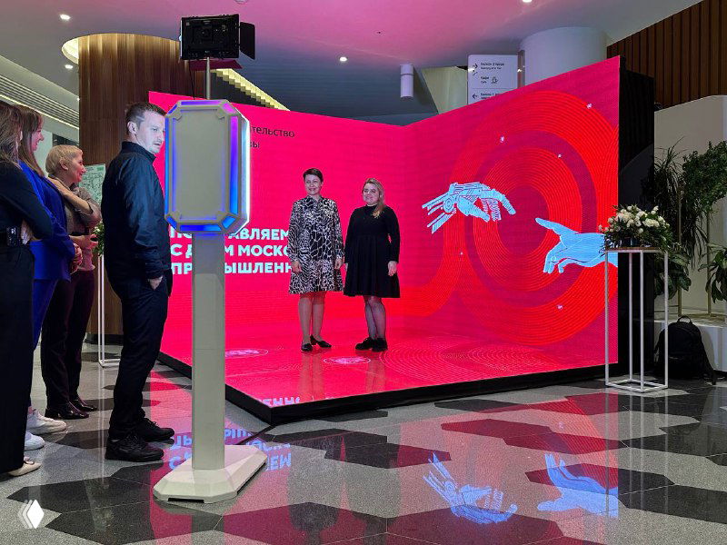 Полукруглый большой LED‑экран на сцене с красочной визуализацией; на переднем плане отдельно стоящая GIF‑фотостойка и отражение на глянцевом полу.