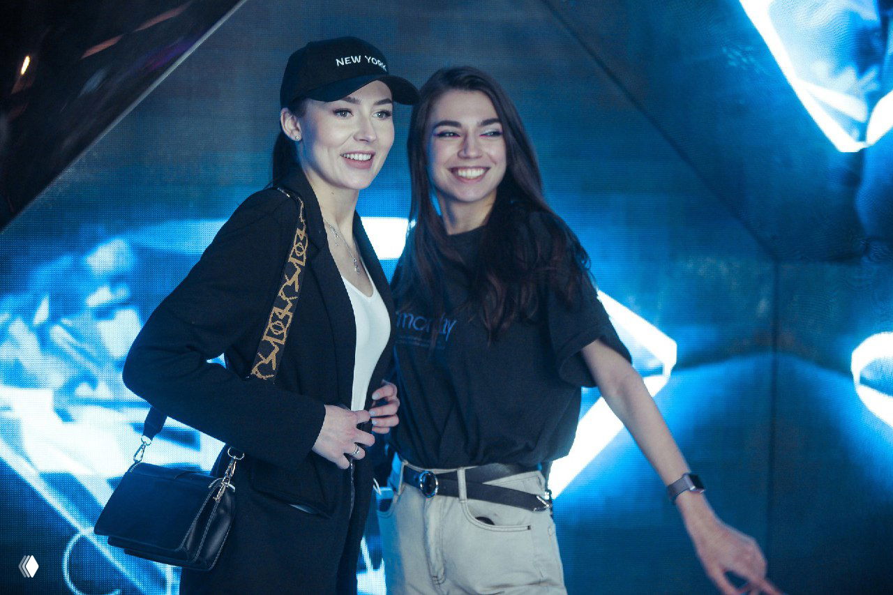 Две женщины улыбаются на фоне LED‑экрана с калейдоскопическим отражением и зеркалами — интерактивная фотозона на мероприятии.