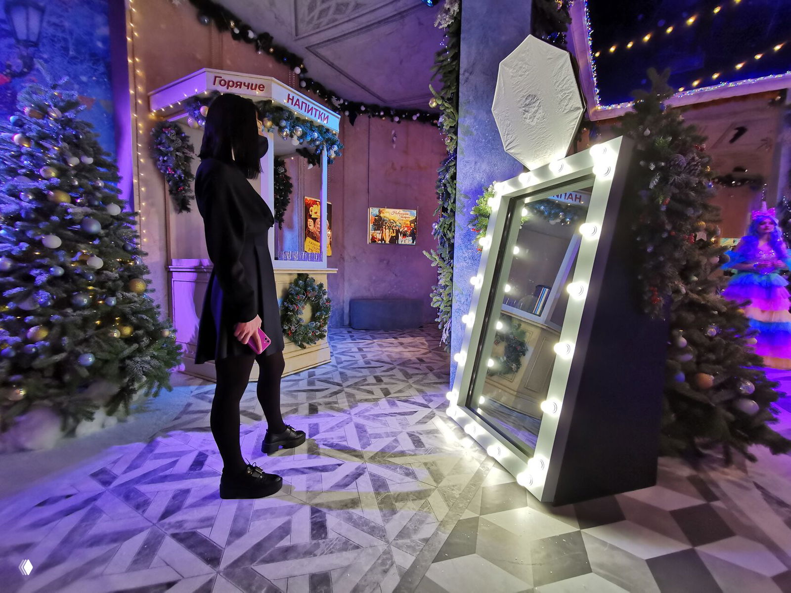 Женщина в тёмном платье стоит напротив подсвеченного селфи-зеркала на праздничной фотозоне с ёлками и украшениями интерьера.