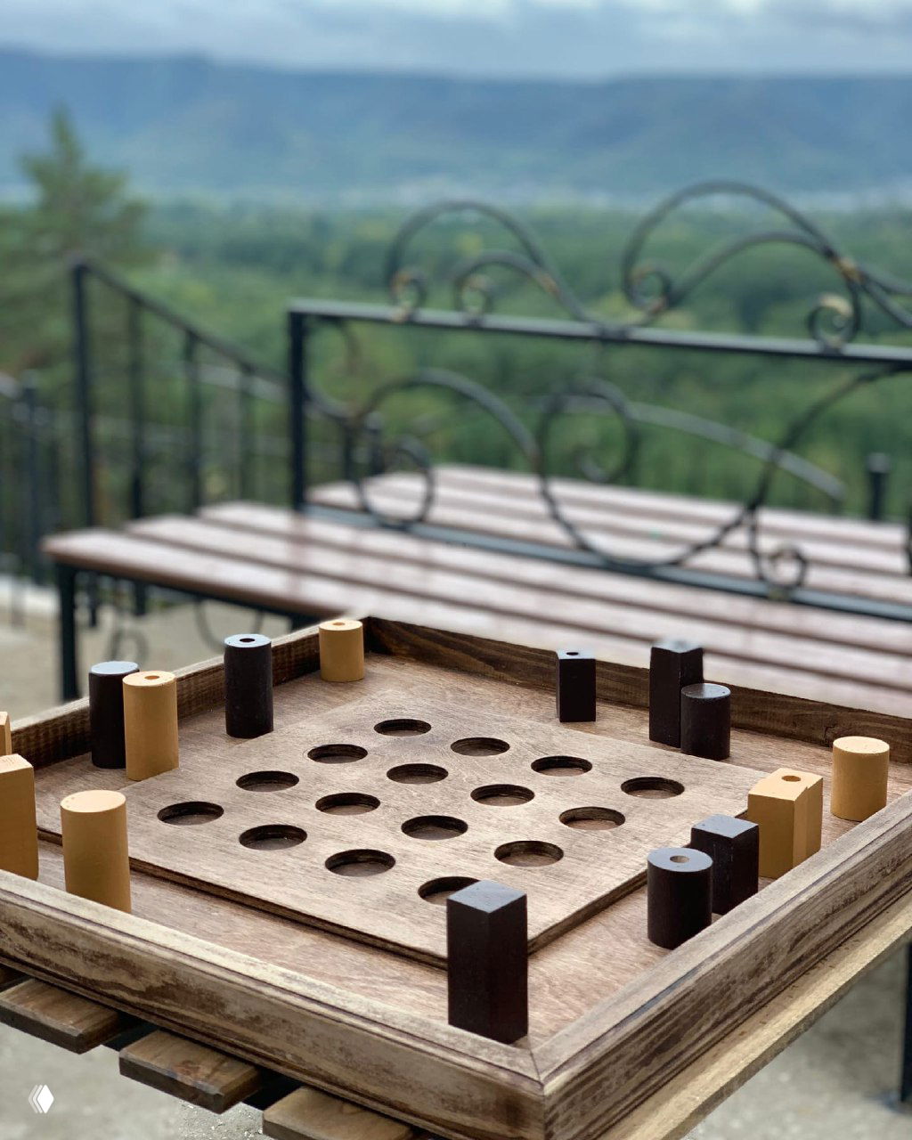 Крафтовая деревянная настольная игра на террасе: игровая доска с круглыми отверстиями и светло‑тёмные фишки, размытый пейзаж