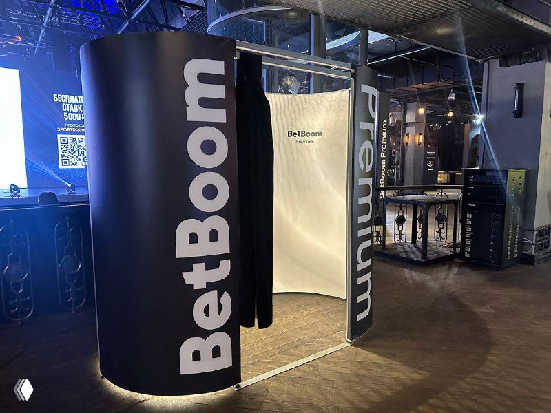 Чёрная классическая фотобудка‑кабина с надписью BetBoom, внутренней светлой шторкой и локальной подсветкой, установленная в зале мероприятия.