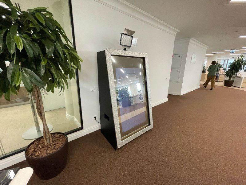 Селфи‑зеркало, стоящее у стены в коридоре: корпус зеркала в металлизированной раме, на экране отражение интерьера и людей на фоне.