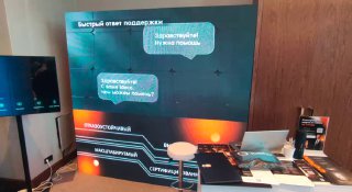 Фотография выставочного стенда с крупным светодиодным LED‑экраном на стене, небольшим столом с брошюрами и промо‑материалами.