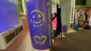 Фиолетовая цилиндрическая фотобудка со шторкой, на корпусе нарисованы смайлик и сердце; человек у входа, интерьер события в помещении.