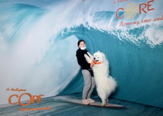 Гости позируют в студийной 3D-фотозоне Bullet Time: человек с большой белой собакой на серфборде на фоне крупной волны в декоре