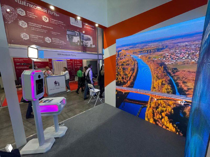 Фотозона на выставке: крупный светодиодный LED‑экран на полу и сзади, в переднем плане белая фотостойка/GIF‑станция с розовой подсветкой и сенсором.