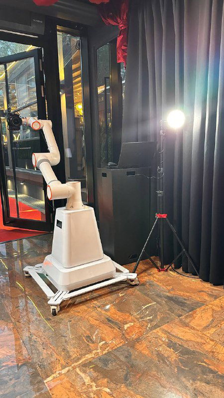 Роботизированная рука с камерой на мобильной платформе в интерьерной фотозоне; видны напольное покрытие, световое оборудование и чёрные шторы вокруг зоны.