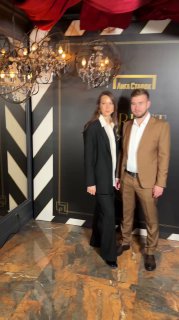 Двое гостей позируют у бренд‑фона в вечерней одежде в зоне съёмки; на заднем плане люстра и декоративные элементы фотозоны.