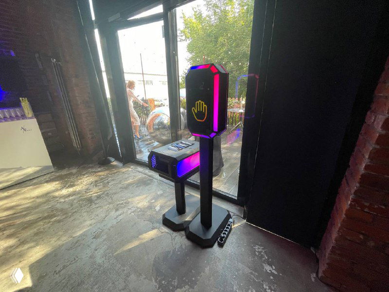 Вертикальная сенсорная стойка TECH‑SCAN с подсветкой и иконкой ладони, установленная у входа на ивент‑локации, вид со стороны.