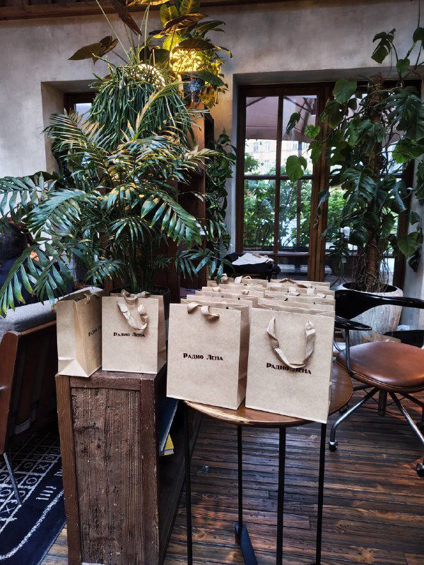Стол с крафтовыми подарочными пакетами на фоне большого количества зелёных растений в уютной локации с деревянным полом — реквизит для мероприятия.