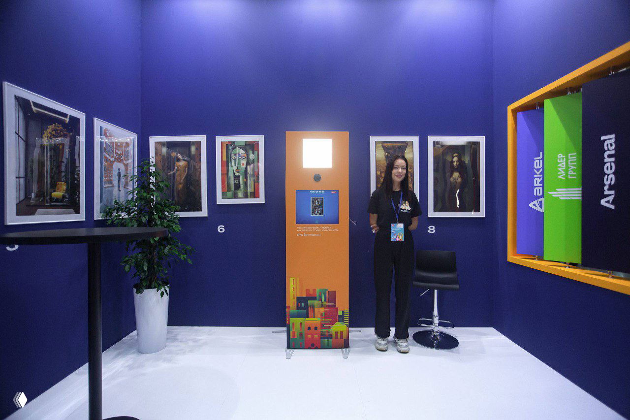 Фотостойка на белом полу выставки: сотрудница рядом, вокруг висят оформленные фотоработы и элементы оформления стенда, демонстрация размещения сервиса.