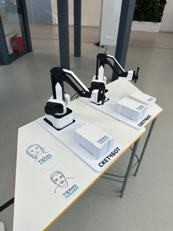 Стол с двумя роботизированными манипуляторами, рядом бумажные наброски портретов и подписи «СКЕТЧБОТ», видно выставочную студию и образцы штриховых рисунков.