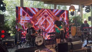 Группа на сцене перед большим светодиодным экраном: барабаны, перкуссия и усилители, яркая графика на заднике для концерта или ивента.