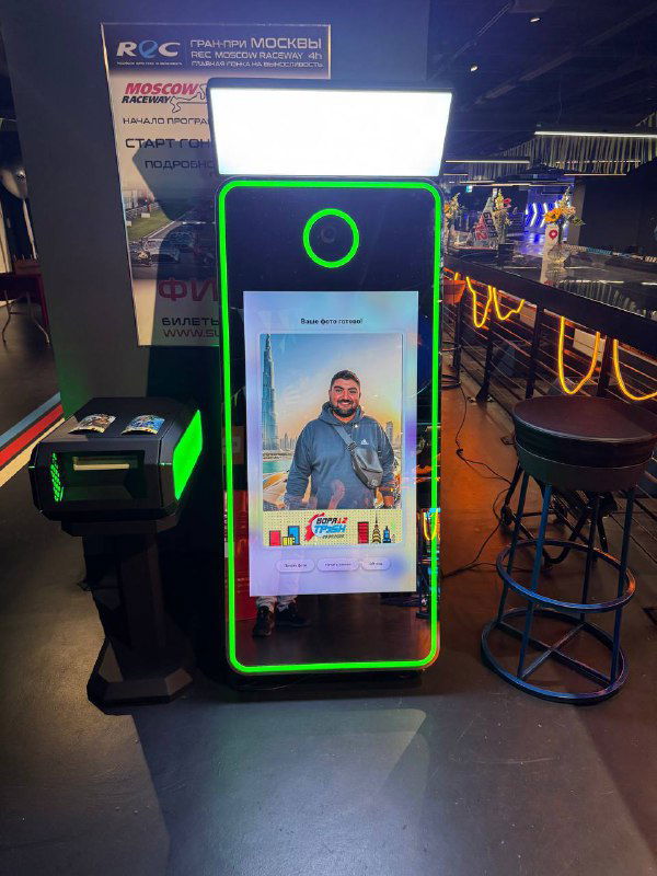Селфи‑зеркало Hi‑Tech в действии: на экране виден пример снимка гостя, яркая зелёная LED‑рамка и стойка с принтером слева для сувенирных фотографий.