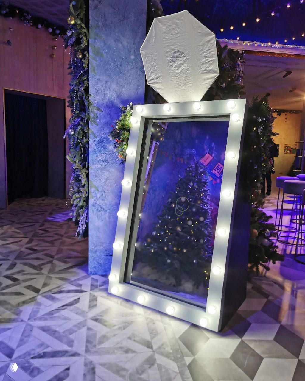Селфи-зеркало в белой рамке с лампочками на фоне праздничной ёлки и голубо‑фиолетового светового оформления интерьера.