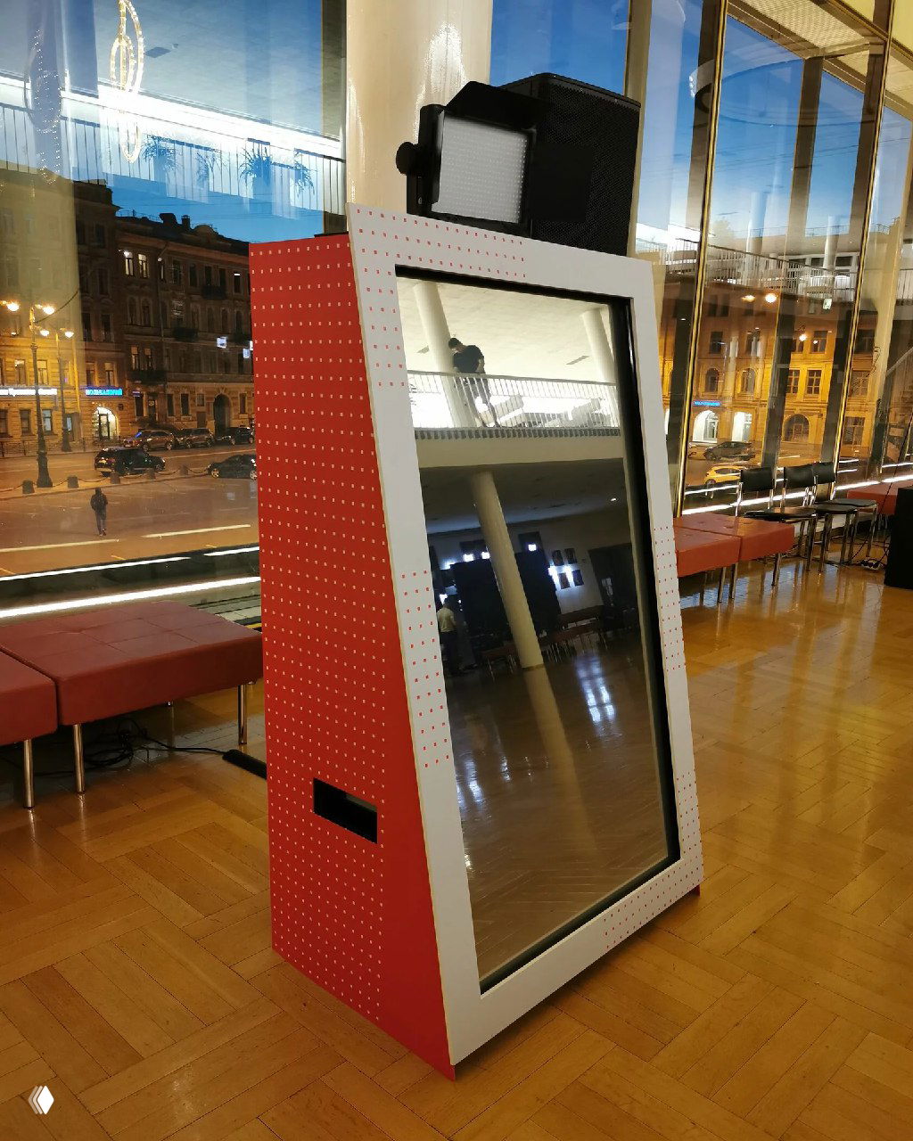 Красно‑белое передвижное селфи‑зеркало в зале у панорамных окон с отражением городского пейзажа и просторного интерьера.