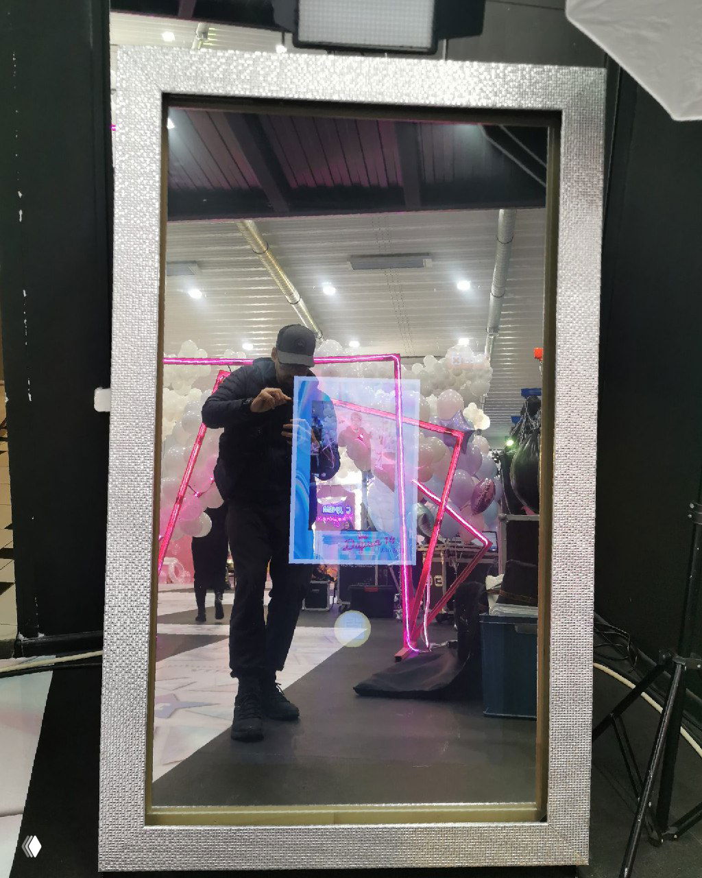 Отражение человека в интерактивном фотозеркале с неоновой подсветкой и шариками на фоне выставочной площадки мероприятия.