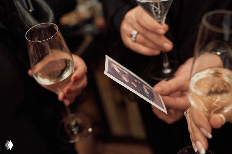 Руки гостей с бокалами шампанского держат моментальную распечатанную фотографию на фуршете — праздничная атмосфера и обсуждение снимка.