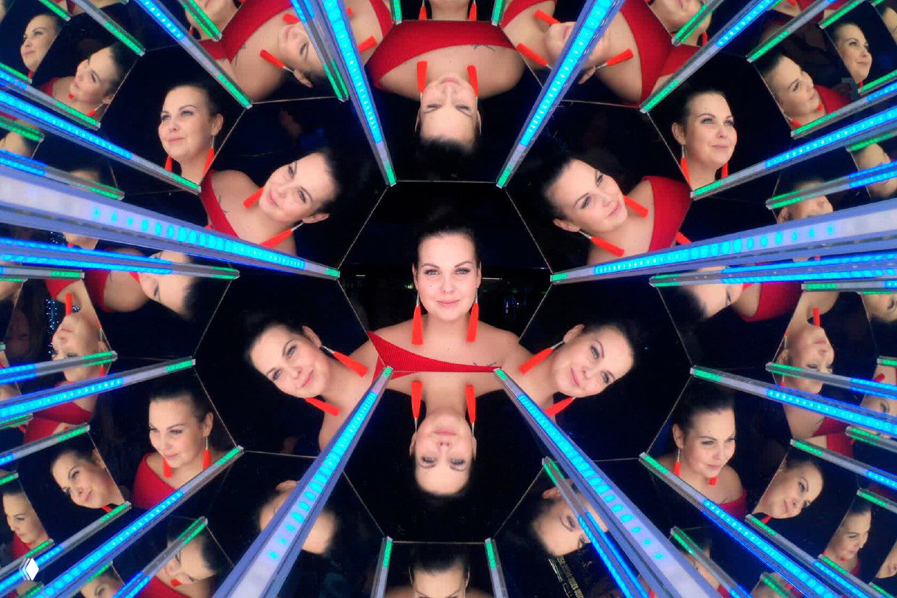 Женщина внутри калейдоскопической фотостойки: множественные зеркальные отражения лица, видны голубые светодиодные полосы.