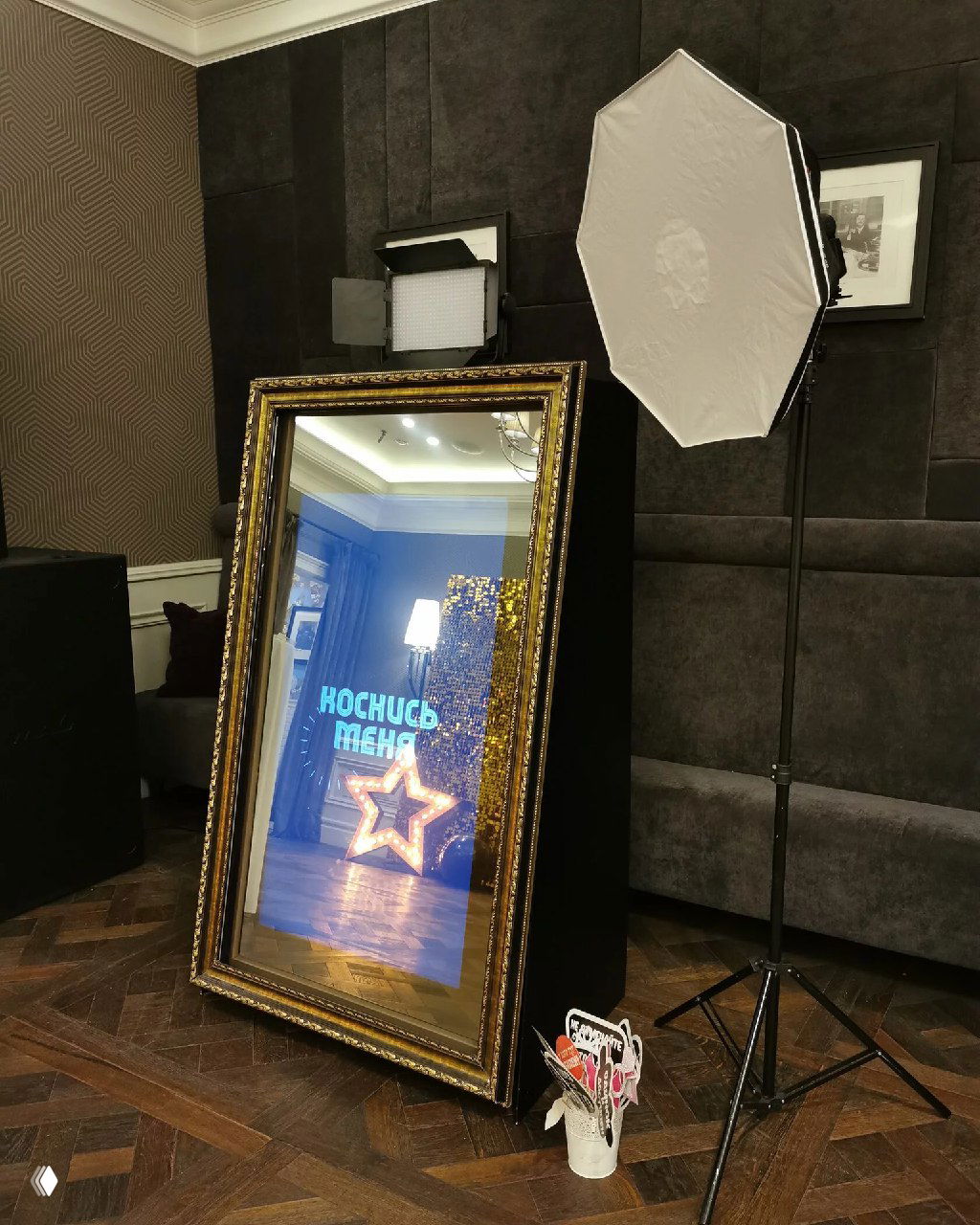 Селфи‑зеркало в золочёной раме в интерьере: большой сенсорный экран, отражение комнаты, студийная подсветка и реквизит рядом.
