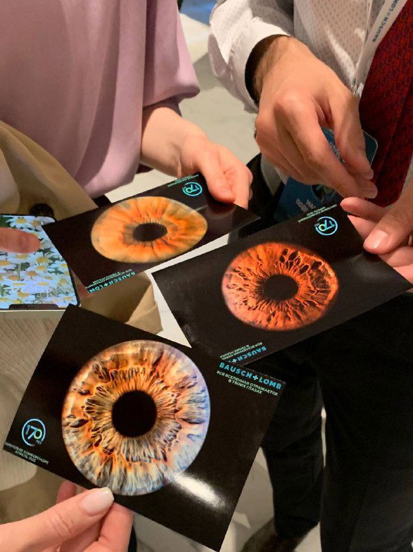 Несколько людей держат напечатанные крупные снимки радужки глаза на чёрных карточках — демонстрация печатной фотопродукции и примеров работ для мероприятия.