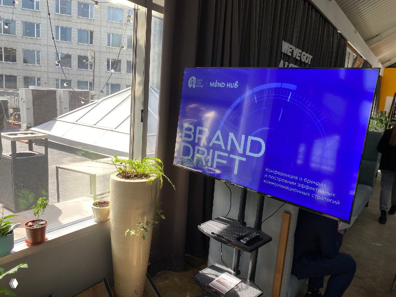 Монитор с заставкой «BRAND DRIFT» у окна в зоне мероприятия: экран, растения на подоконнике и часть интерьера площадки для фотозоны.