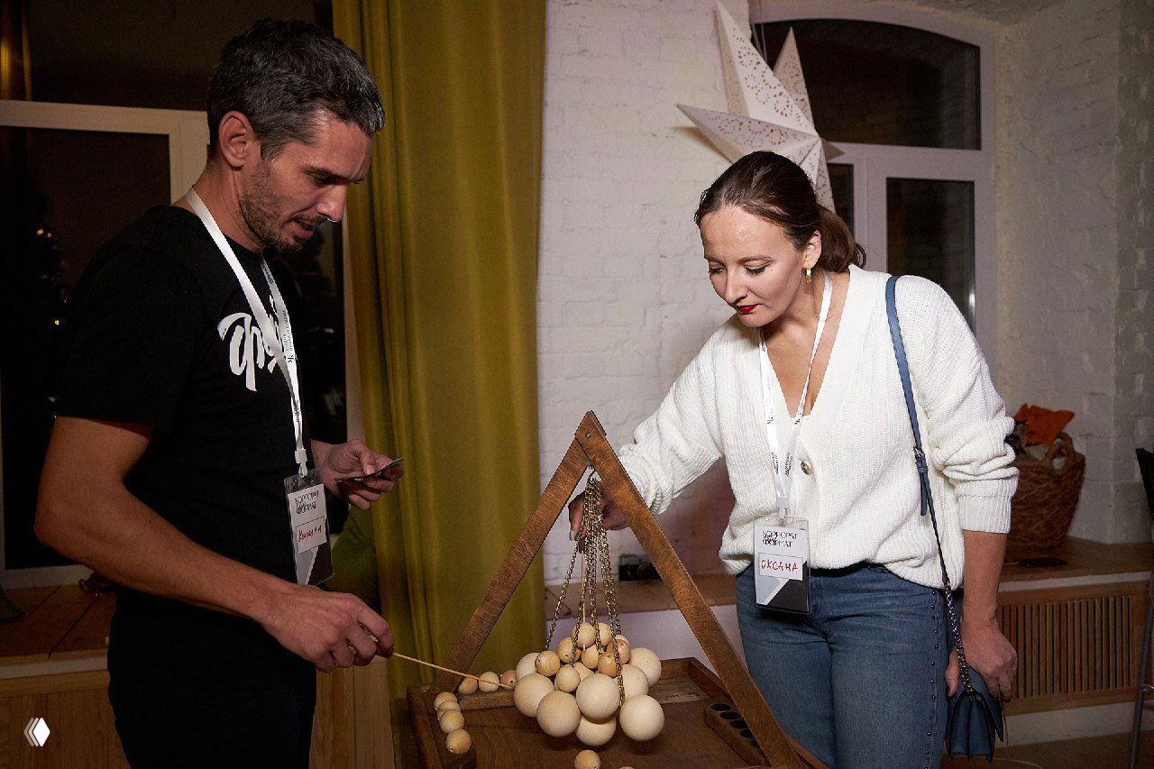 Двое взрослых взаимодействуют с деревянной настольной игрой: шарики на подвесах и рамка, интерактивный реквизит на вечернем мероприятии в помещении.
