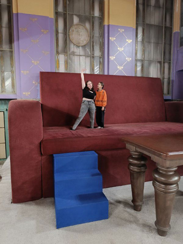 Фото на фоне гигантского дивана из квеста: две девушки (автор и младшая сестра) в миниатюрном масштабе на фоне реквизита, улыбки и настроение.