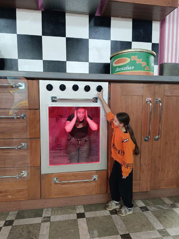 Сцена из квеста у игрушечной кухни: девочка открывает духовку, а взрослая рядом реагирует — забавный момент семейной вылазки и интерактивных сцен.