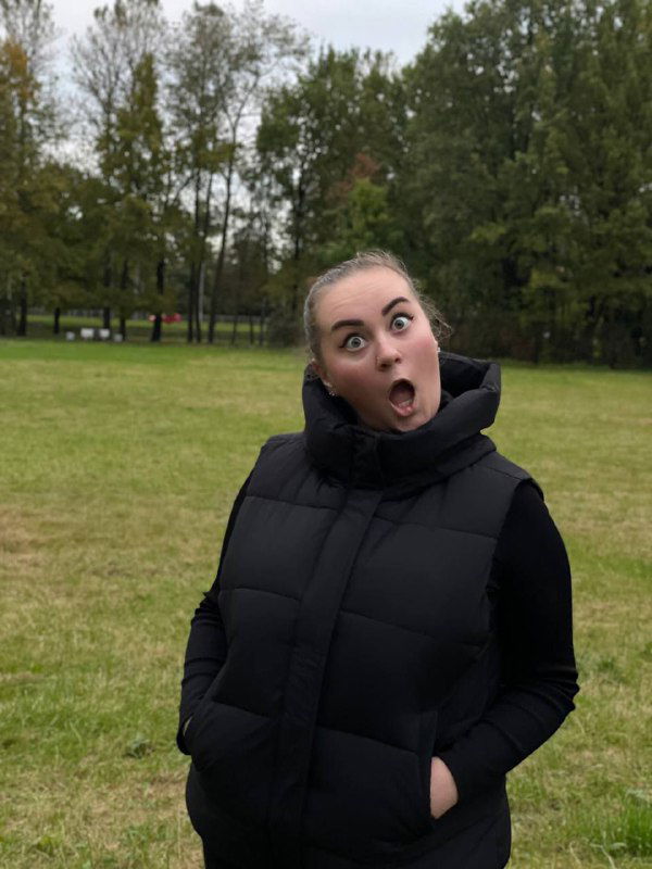 Та же женщина в тёмном жилете на газоне делает удивлённое лицо в парке среди деревьев; кадр подчёркивает искренность и игровую сторону автора в блоге.