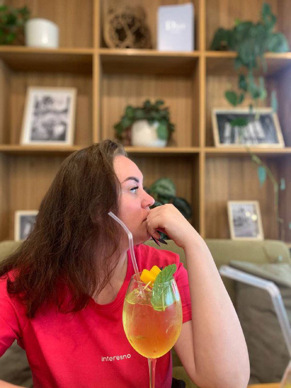 Портрет девушки в кафе с ярким апельсиновым коктейлем в руке: тёплый интерьер, деревянные полки и задумчивый взгляд в сторону окна.