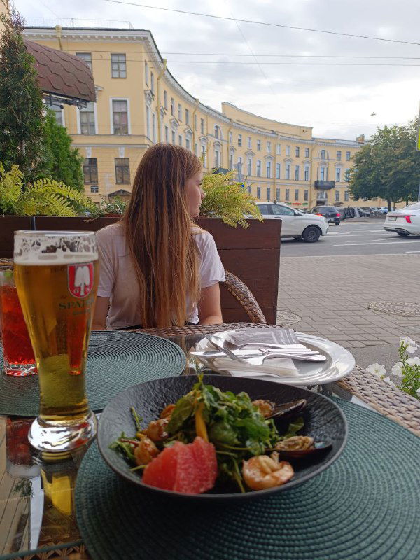 Салат и бокал пива на столе, девушка смотрит в сторону улицы: кадр подчёркивает спокойный вечер в городе и моменты вне офиса.