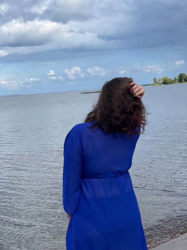 Женщина в синем платье стоит у берега Финского залива, вид сзади: ветер в волосах, обширная водная гладь и лёгкая облачность над горизонтом.