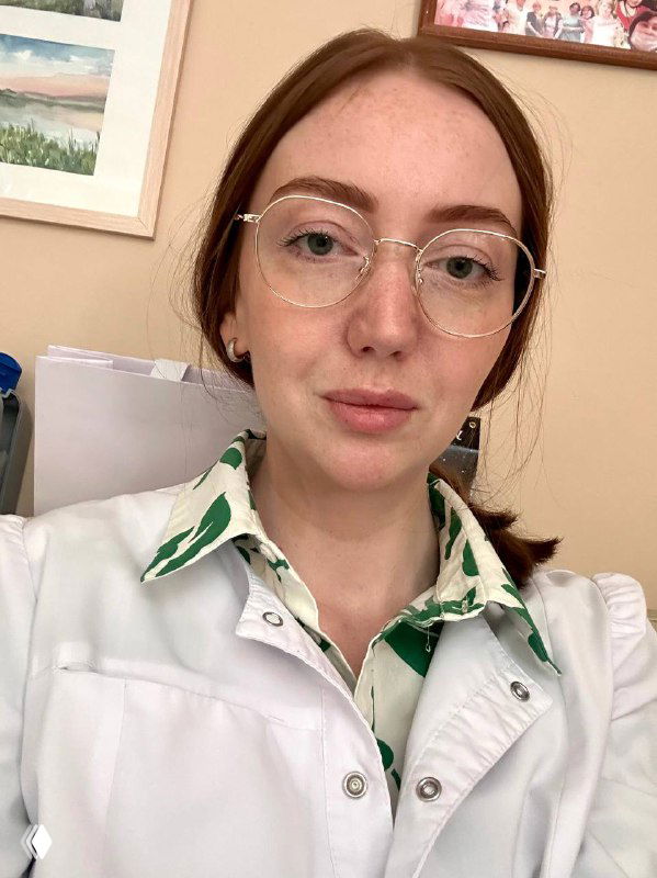 Портрет молодой женщины в белом халате и очках в кабинете клиники — врач-невролог смотрит в камеру.