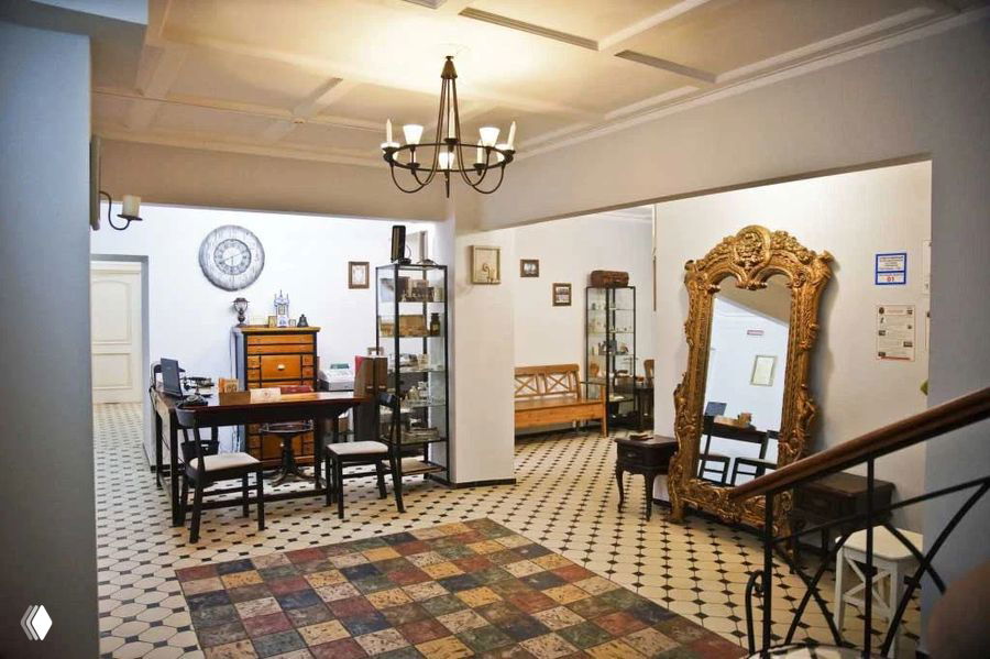Интерьер приёмной «Клиники на Потаповском»: стол и стулья у письменного стола, витрины с предметами, большое резное зеркало в золотой раме и узорный плиточный пол.