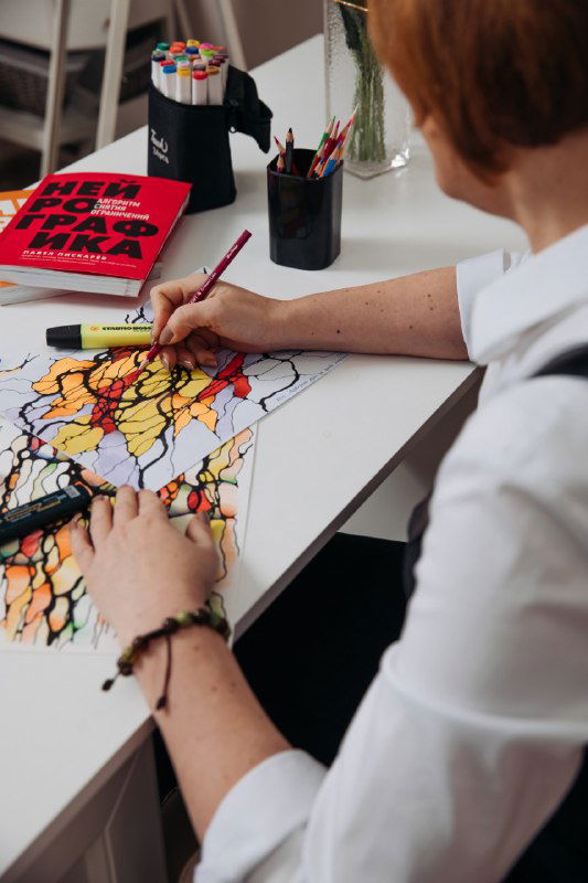 Рука автора за столом дорисовывает нейрографический узор маркером; видны стакан с цветными карандашами, книга и несколько готовых рисунков.