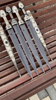 Ракурс сверху на ряд кинжалов: отчетливо виден узорный дамаск клинков и инкрустированные мельхиоровые ножны с разноцветными вставками‑камнями.