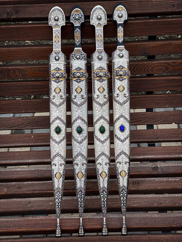 Вид со стороны ножен: четыре ножна с разноцветными вставками‑камнями и тонкой гравировкой на мельхиоре; задний фон — деревянные планки и мощение.