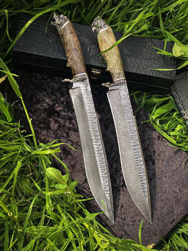Два ножа в целом кадре: сбоку видны клинки с дамасковым рисунком и рукояти из светлой и тёмной карельской березы на траве.