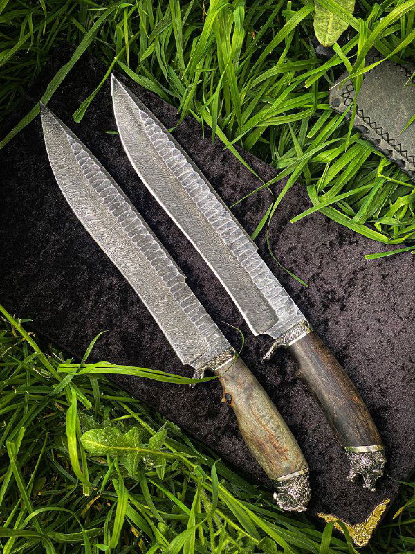 Крупный план двух дамасковых клинков ножей «Медведь/Сова», видна текстура стали и профиль обуха на травяном фоне.