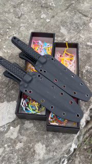 Черные кайдекс-ножны, уложенные на бетонной поверхности рядом с коробками и упаковочной стружкой; видны отверстия для фурнитуры.