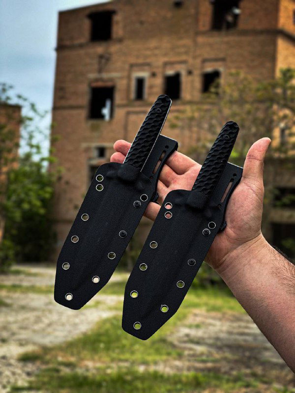 Руки держат черные кайдекс-ножны на фоне полуразрушенного здания; видны заклепки и конструктивные отверстия ножен.