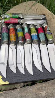 Широкий ракурс нескольких изготовленных ножей с полированными лезвиями и цветными ручками; кадр на фоне травы и дерева, видно оформление ножен и узор гард