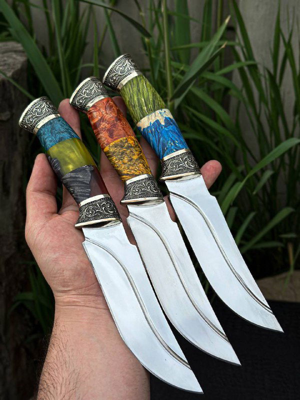 Крупный план трёх ножей в руке мастера: цветные комбинированные рукояти из карельской берёзы и акрила, мельхиоровые гарды и блестящие полированные лезвия из стали 95х18